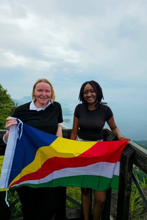 Two smiling women holding a colorful flag with blue, yellow, red, and green stripes on a mountain overlook with cloudy sky background