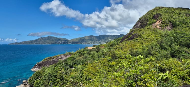 Aerial view of a lush green hillside overlooking turquoise tropical waters and distant mountains under blue sky with clouds