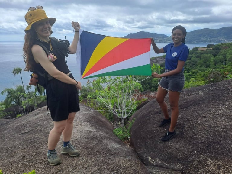 Two people holding the Seychellois flag on a rocky hilltop overlooking ocean and mountains