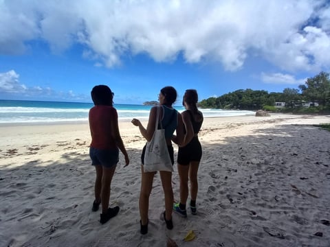 Three people standing on a sandy beach facing the ocean under a blue sky with white clouds