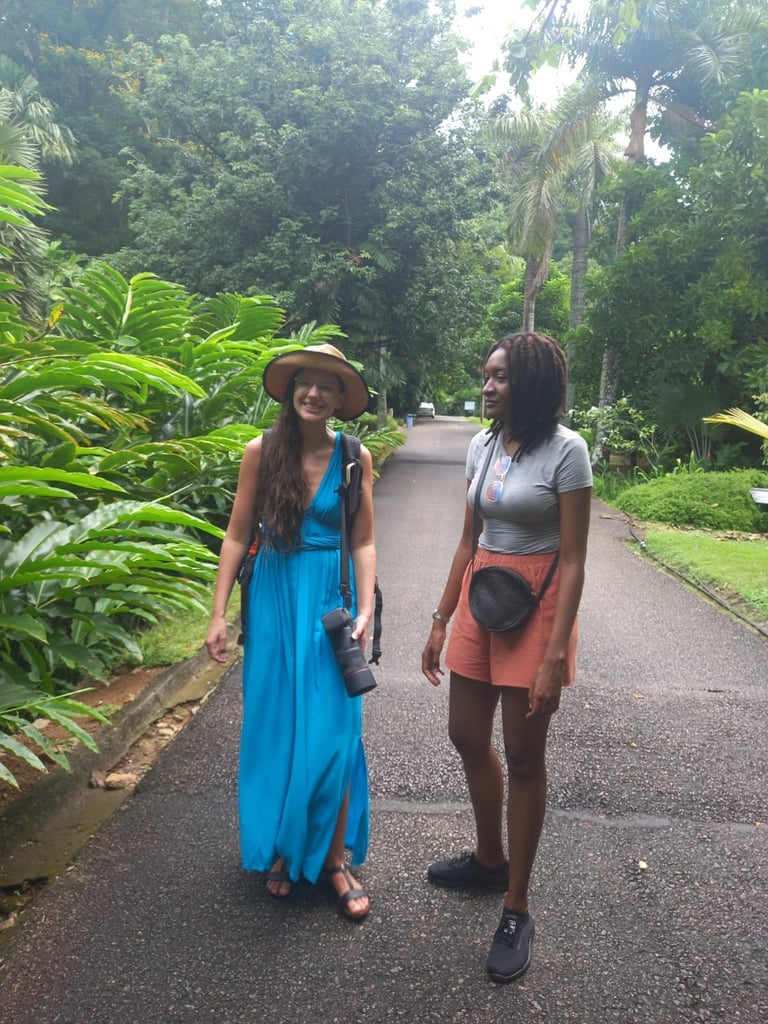 Two women walking on a tree-lined path, one wearing a blue dress and hat, the other in a gray shirt and orange skirt