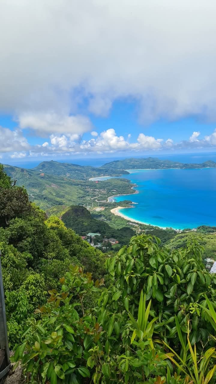 Elevated tropical coastal landscape with turquoise ocean, lush green mountains, white sand beaches, and blue sky with white clouds