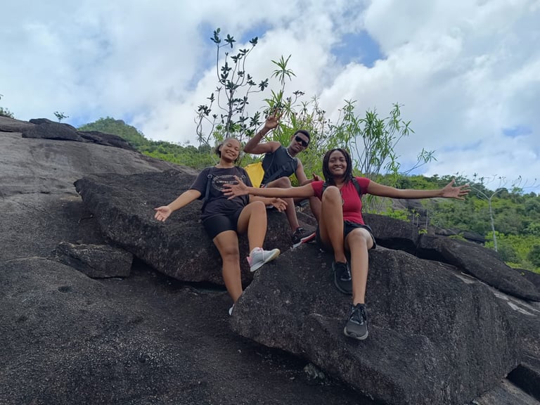 Three people sitting on large boulders outdoors on a hillside with lush green vegetation and cloudy sky